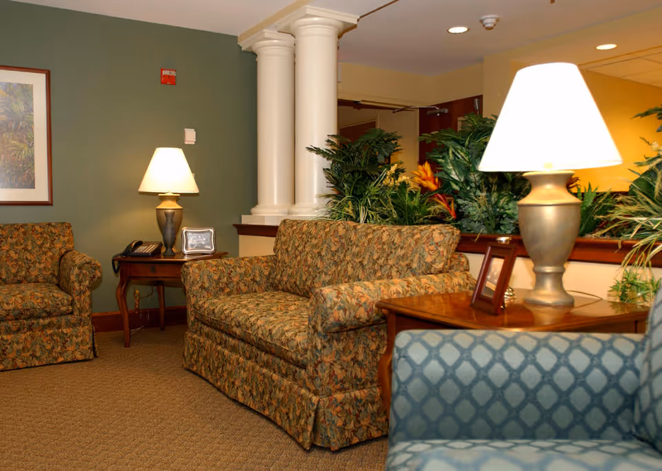 A cozy seating area in a senior living facility with patterned upholstered armchairs and a loveseat, two wooden side tables with lamps, framed pictures, and lush green plants in the background. The walls are painted green and beige, and there are two white decorative columns near the center.