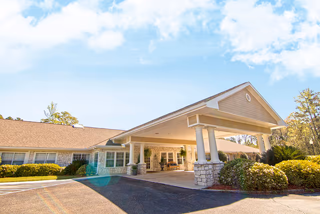 Exterior view of a single-story building with a covered entrance supported by white columns, surrounded by bushes and trees under a partly cloudy sky.