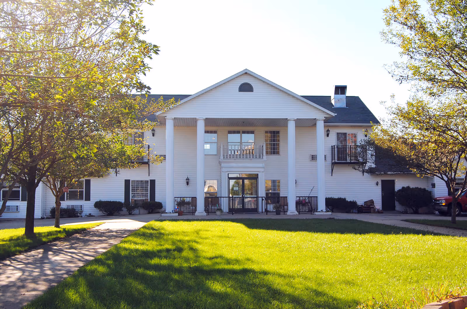 Front exterior view of a two-story white building with large white columns supporting a triangular pediment above the entrance. The building is surrounded by green grass and trees with sunlight casting shadows on the lawn.