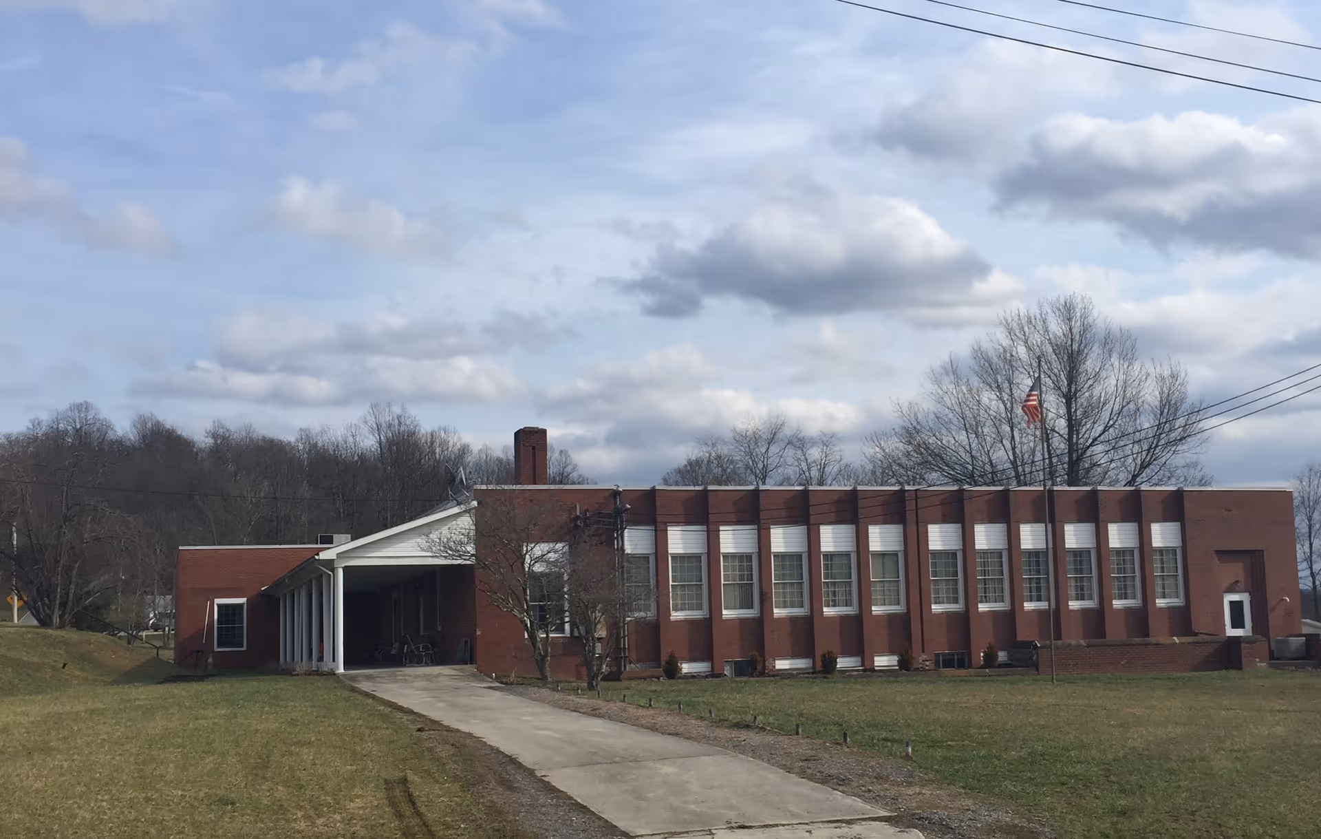 Exterior view of a single-story brick building with multiple windows and a covered entrance. The building is surrounded by grass and trees with a cloudy sky overhead. An American flag is visible on a flagpole near the building.