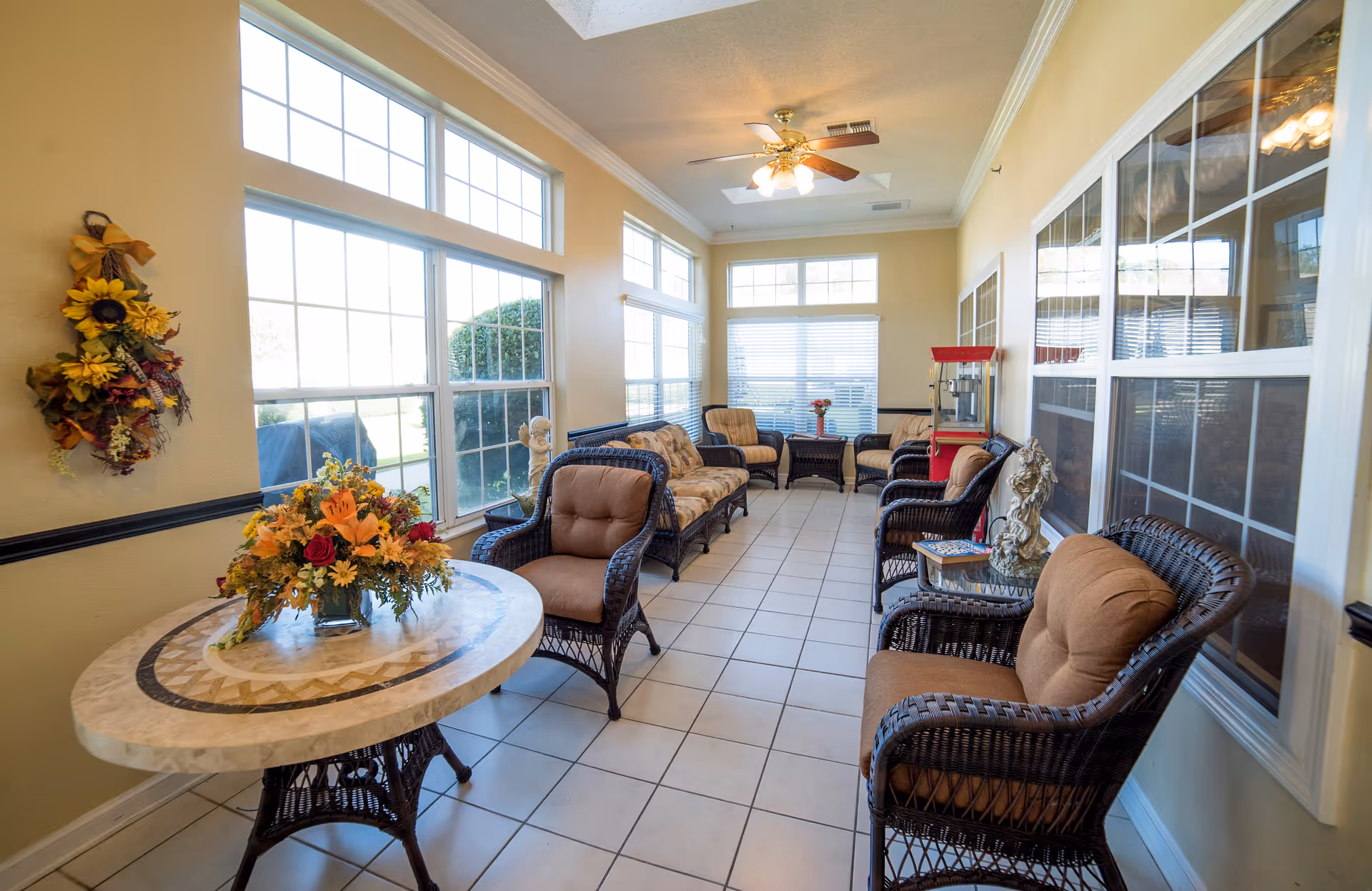 A bright sunroom with large windows on two sides letting in natural light. The room features wicker furniture with brown cushions, including chairs and a sofa. There is a round table with a floral arrangement on it, a ceiling fan with lights, and a red popcorn machine in the corner. The walls are painted light yellow with white trim and tile flooring.