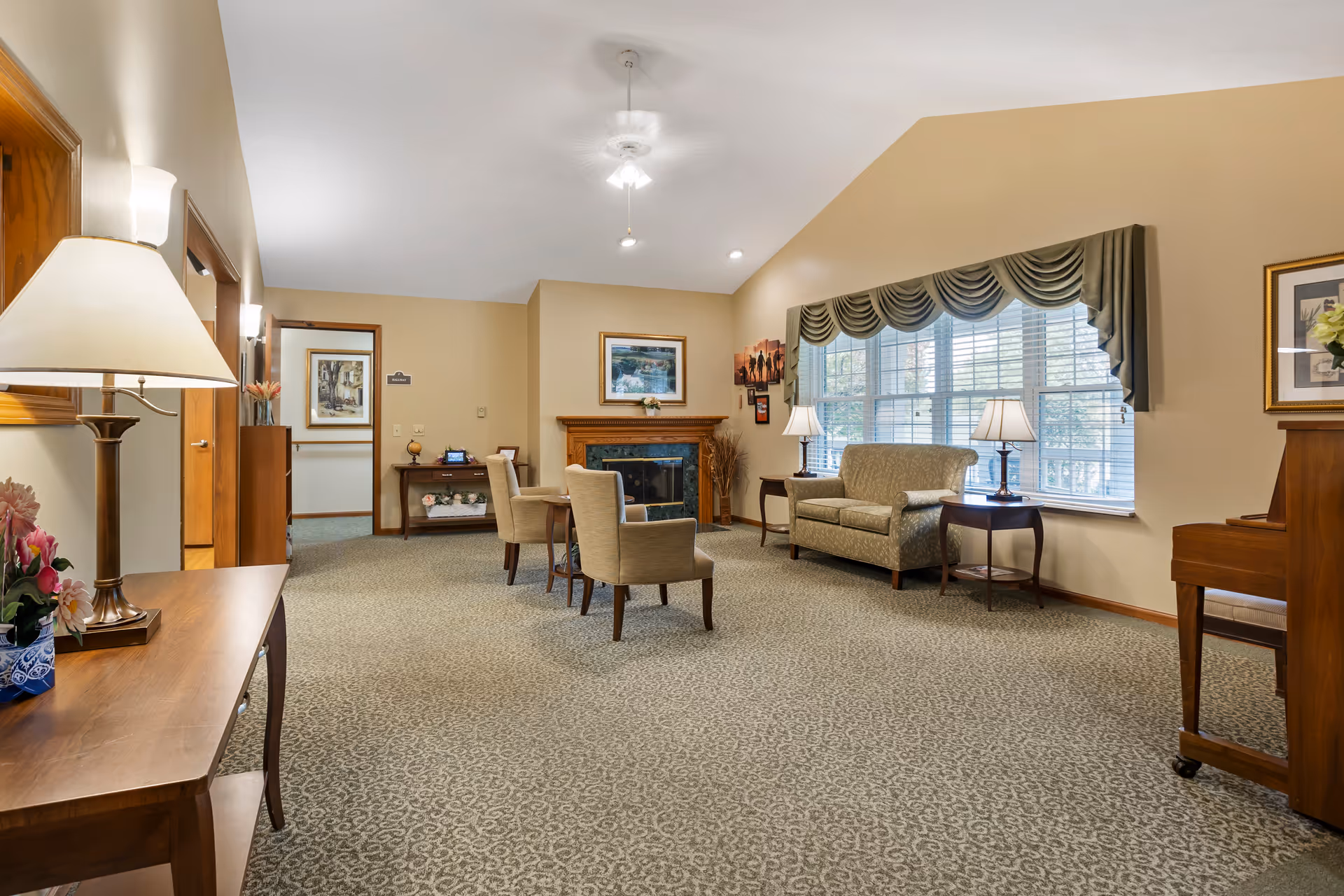 A bright communal living room with chairs, a loveseat, side tables and lamps, a fireplace, and a large window with a valanced curtain.