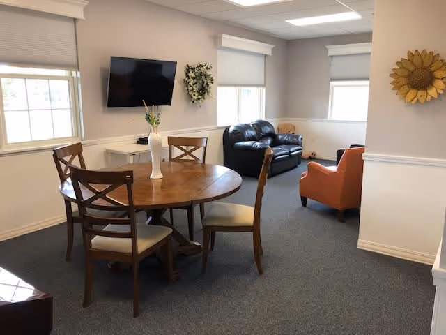 A cozy common area with a round wooden table and four chairs in the foreground. A white vase with flowers is on the table. In the background, there is a black leather loveseat and an orange armchair near two windows with white blinds. A flat-screen TV is mounted on the wall above a small white cabinet, and a decorative wreath hangs beside it. The room has light-colored walls and carpeted flooring.