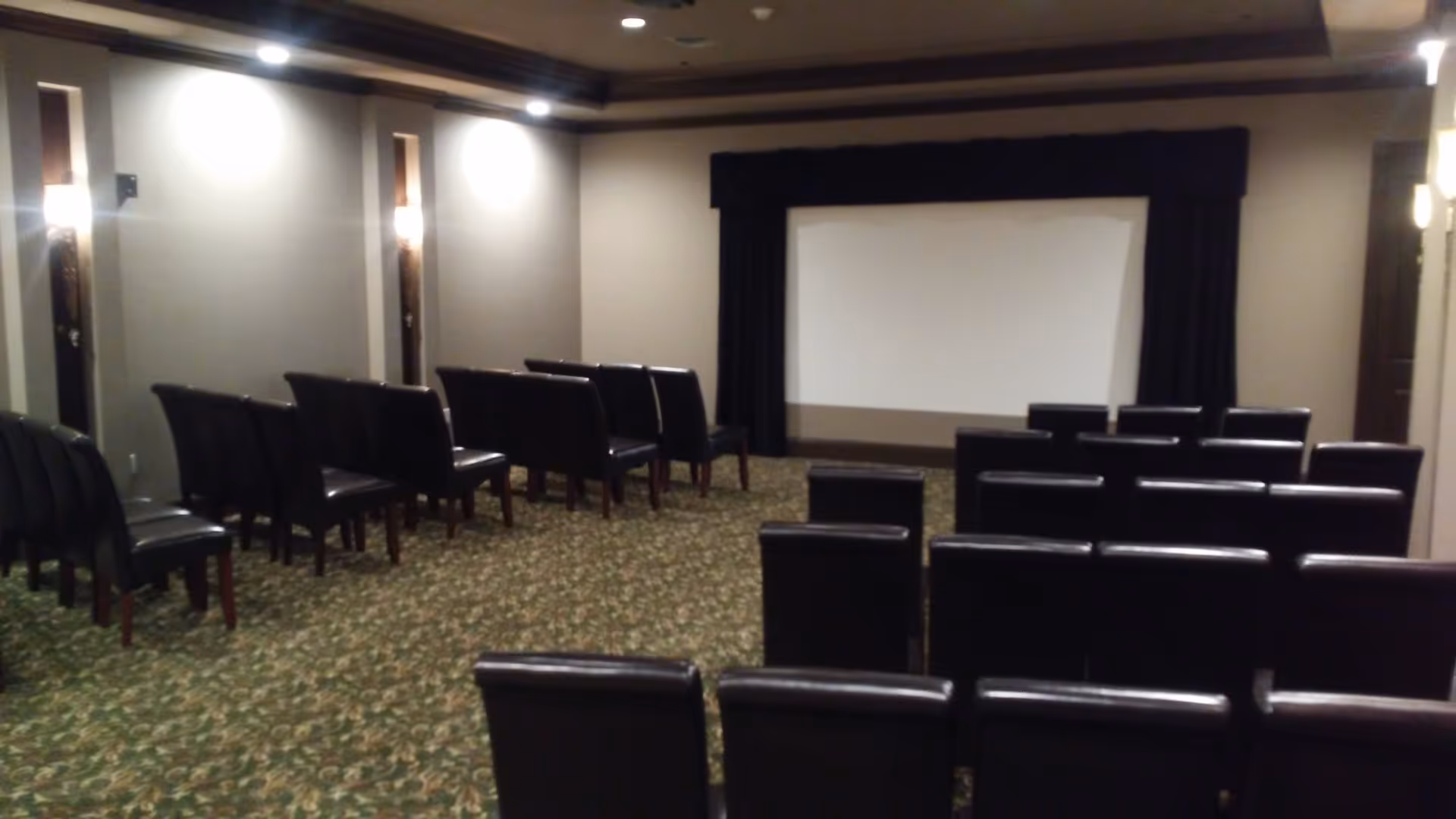 A small theater room with rows of black cushioned chairs facing a large white projection screen framed by dark curtains. The room has beige walls with wall sconces providing soft lighting and a patterned carpet floor.