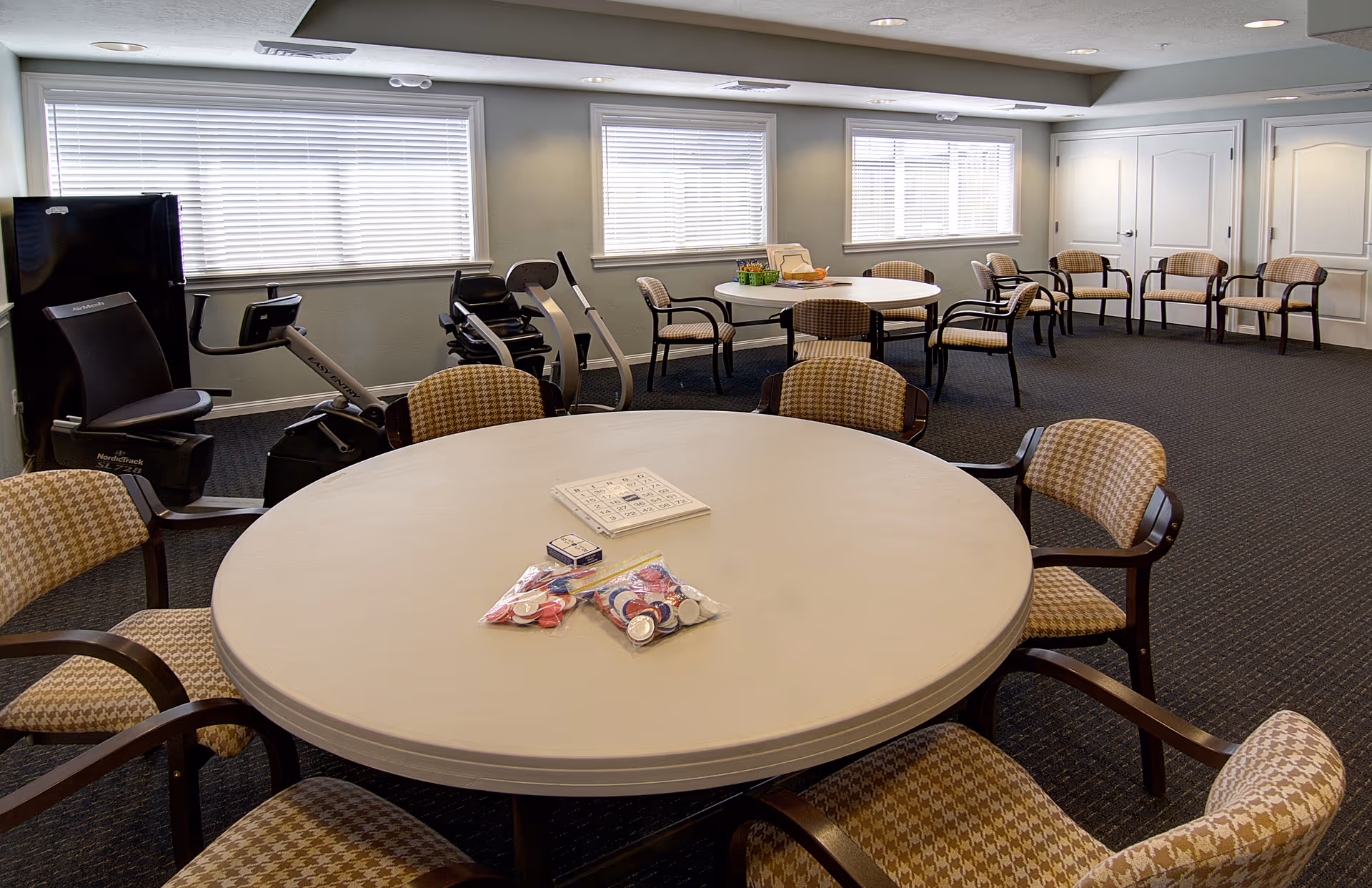 A spacious room with round tables surrounded by cushioned chairs with a houndstooth pattern. On the tables are some game pieces and a bingo card. In the background, there are exercise machines near windows with blinds, and the room has neutral-colored walls and carpeted flooring.