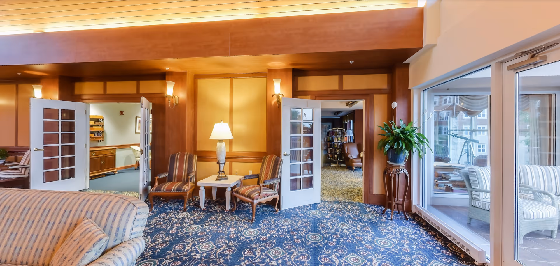 A cozy interior common area in a senior living facility with patterned blue carpet, striped upholstered chairs, a small table with a lamp, and double glass doors leading to other rooms. There is a large window on the right side showing an adjacent sunroom with additional seating and a telescope.