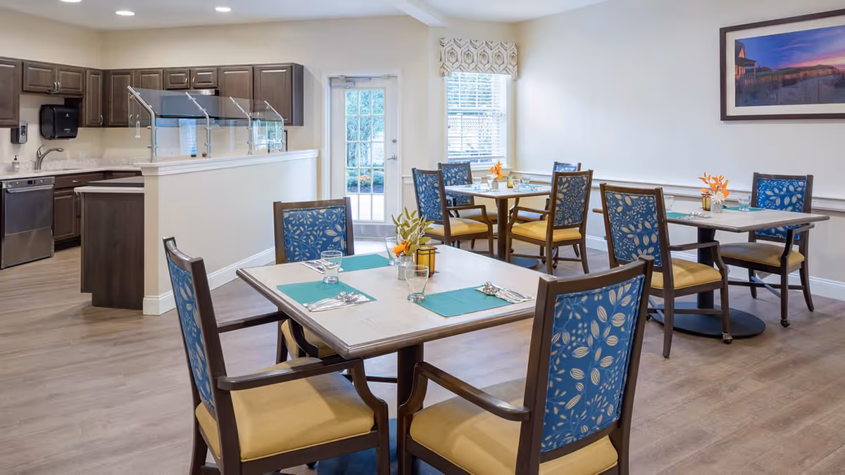 A bright dining area with several square tables set with placemats, glasses, and silverware. Each table is surrounded by wooden chairs with blue patterned backs and yellow cushions. In the background, there is a kitchen area with dark wood cabinets, a dishwasher, and a countertop with glass sneeze guards. A door and window let in natural light, and a framed picture hangs on the wall.