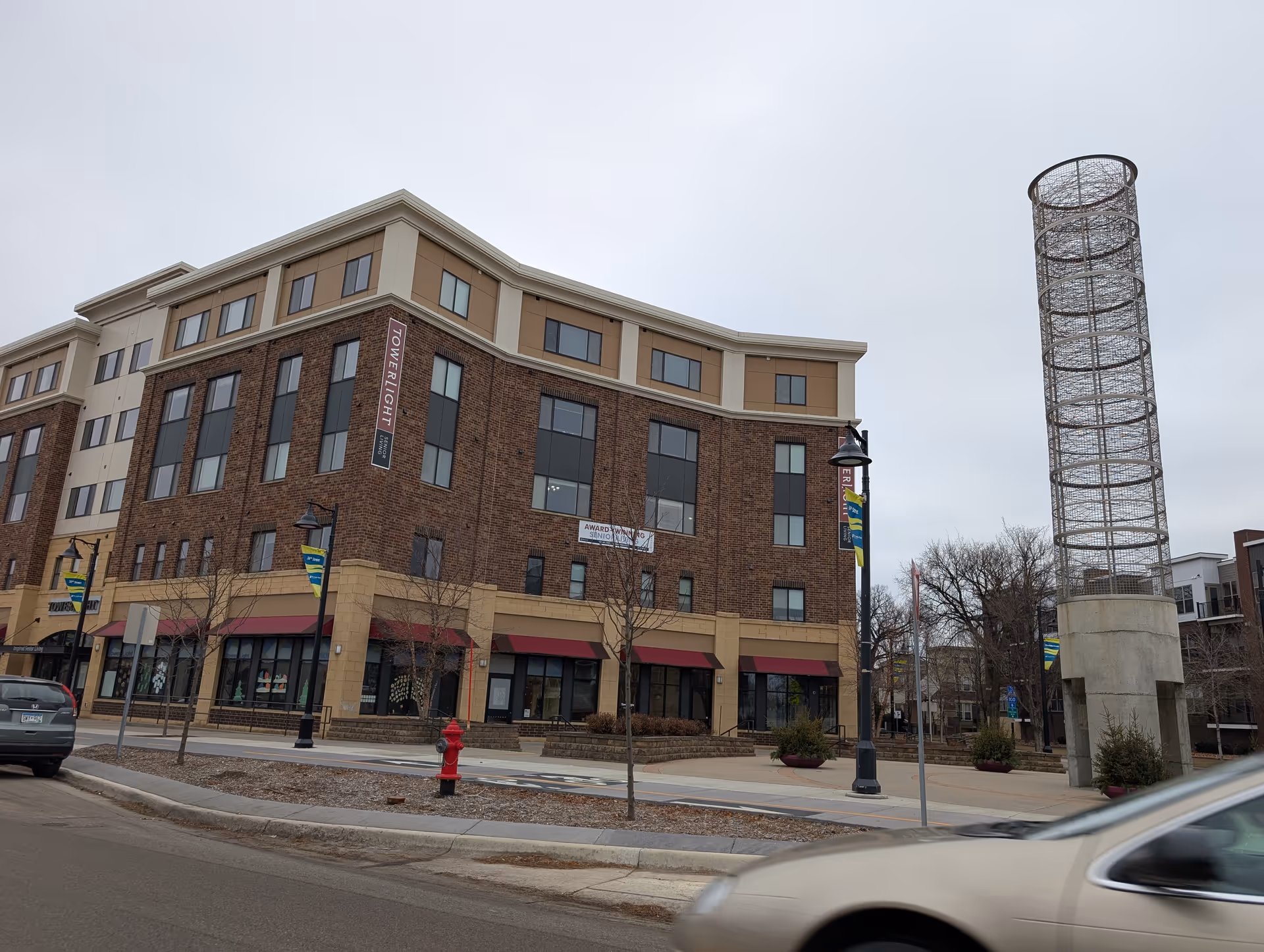 Four-story brick and tan mixed-use building labeled "Towerlight" with storefronts and a tall cylindrical metal sculpture in the plaza on a cloudy day.