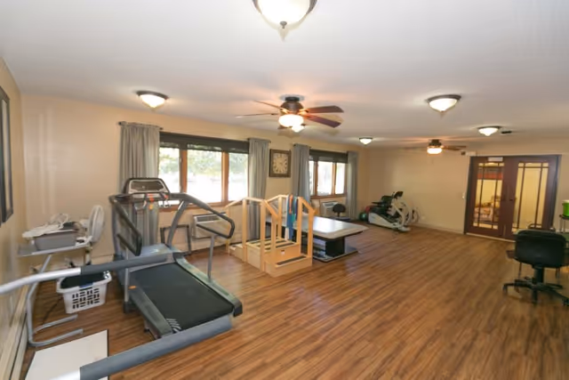 A spacious room with wooden flooring and beige walls, featuring exercise equipment including a treadmill, a stationary bike, and a wooden balance ramp. The room has multiple windows with curtains, ceiling fans with lights, and a clock on the wall. There is also a black office chair and a desk in the corner.