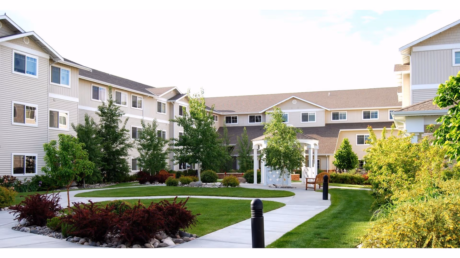 Outdoor courtyard area of a senior living facility with a paved walkway, green grass, various trees and shrubs, benches, and a white gazebo. The building surrounding the courtyard is three stories tall with beige siding and multiple windows.