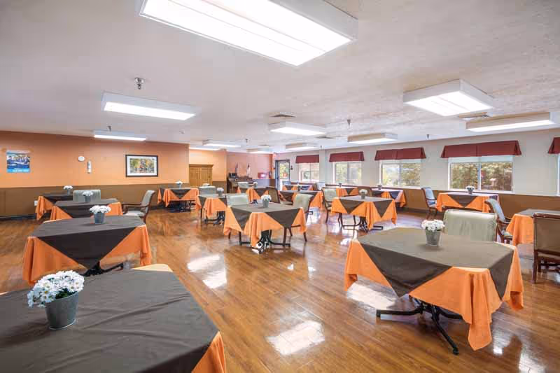 A spacious dining room with multiple tables covered with orange tablecloths and brown overlays, each table decorated with a small potted white flower. The room has large windows with red valances, wooden flooring, and bright overhead lighting.