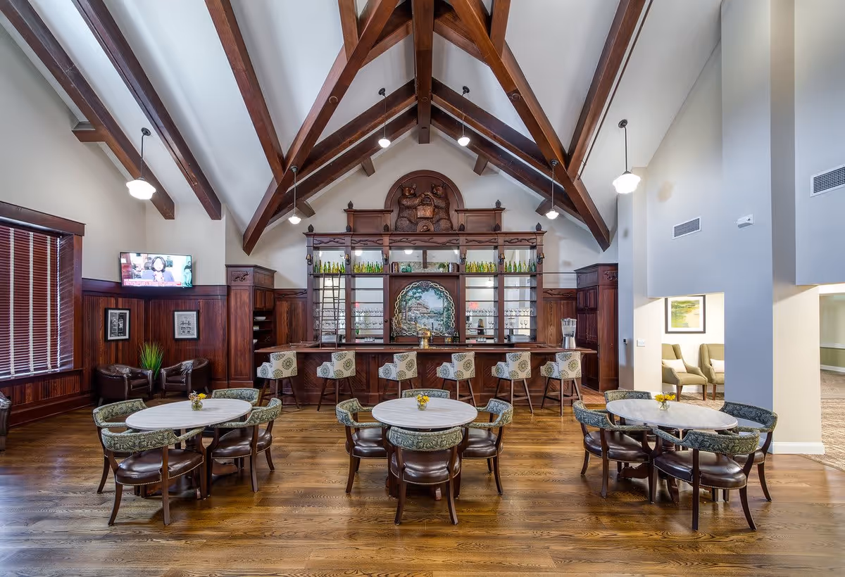 Interior of a senior living facility common area with wooden floors, round tables with chairs, a bar with high stools, and a high vaulted ceiling with exposed wooden beams. There is a TV mounted on the wall to the left and decorative artwork behind the bar.