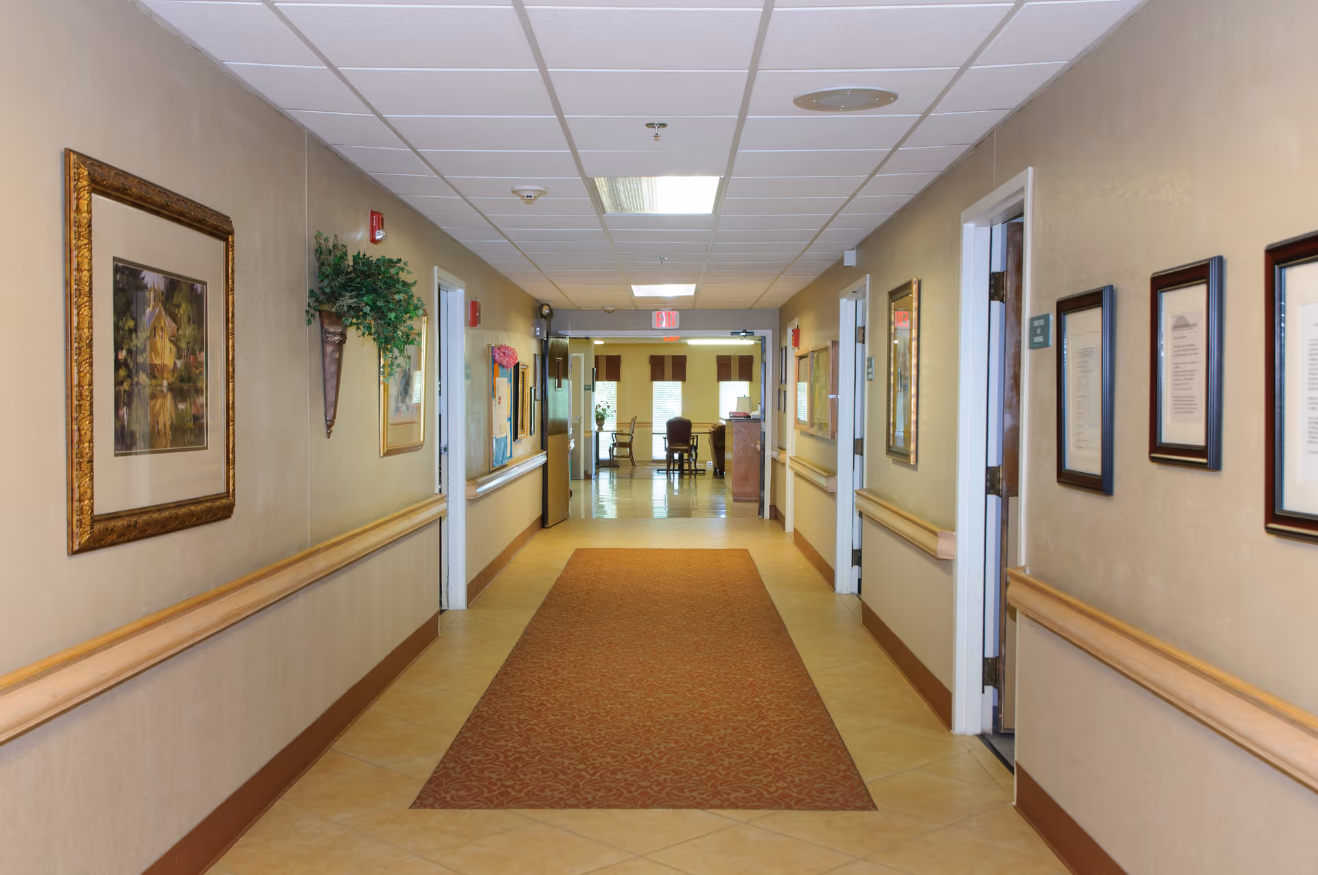 A well-lit hallway in a senior living facility with beige walls, a patterned carpet runner, framed artwork and documents on the walls, handrails on both sides, and several open doorways leading to other rooms. At the end of the hallway, there is a seating area with chairs and windows letting in natural light.