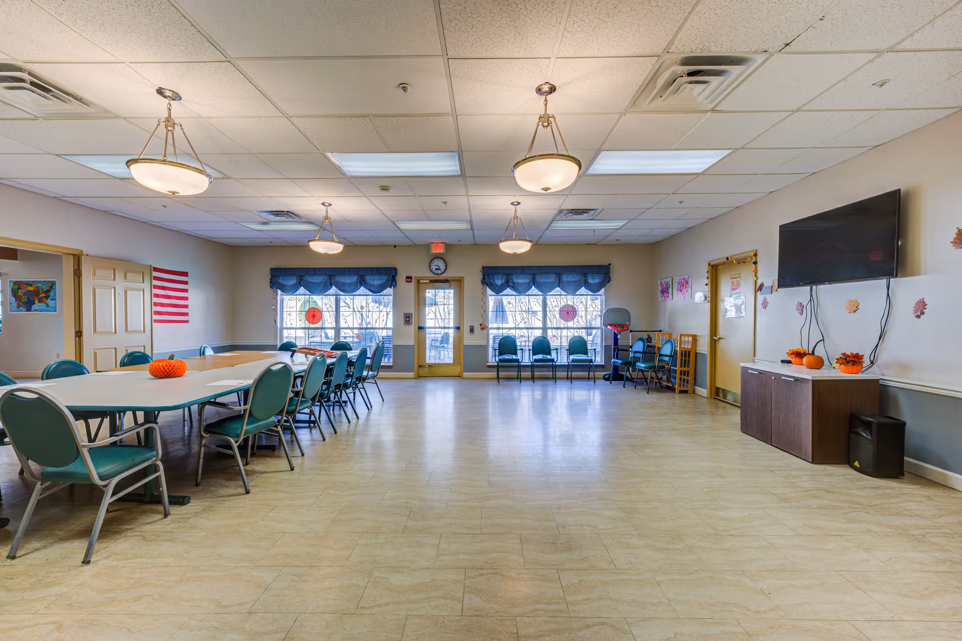 A large, well-lit community room with a long table surrounded by green chairs on the left side. The room has beige walls, a tiled floor, and several ceiling lights. There are two large windows with blue valances and a door in the center back wall. On the right side, there is a mounted flat-screen TV above a cabinet decorated with small pumpkins and fall-themed decorations. Additional chairs are lined up against the back wall.