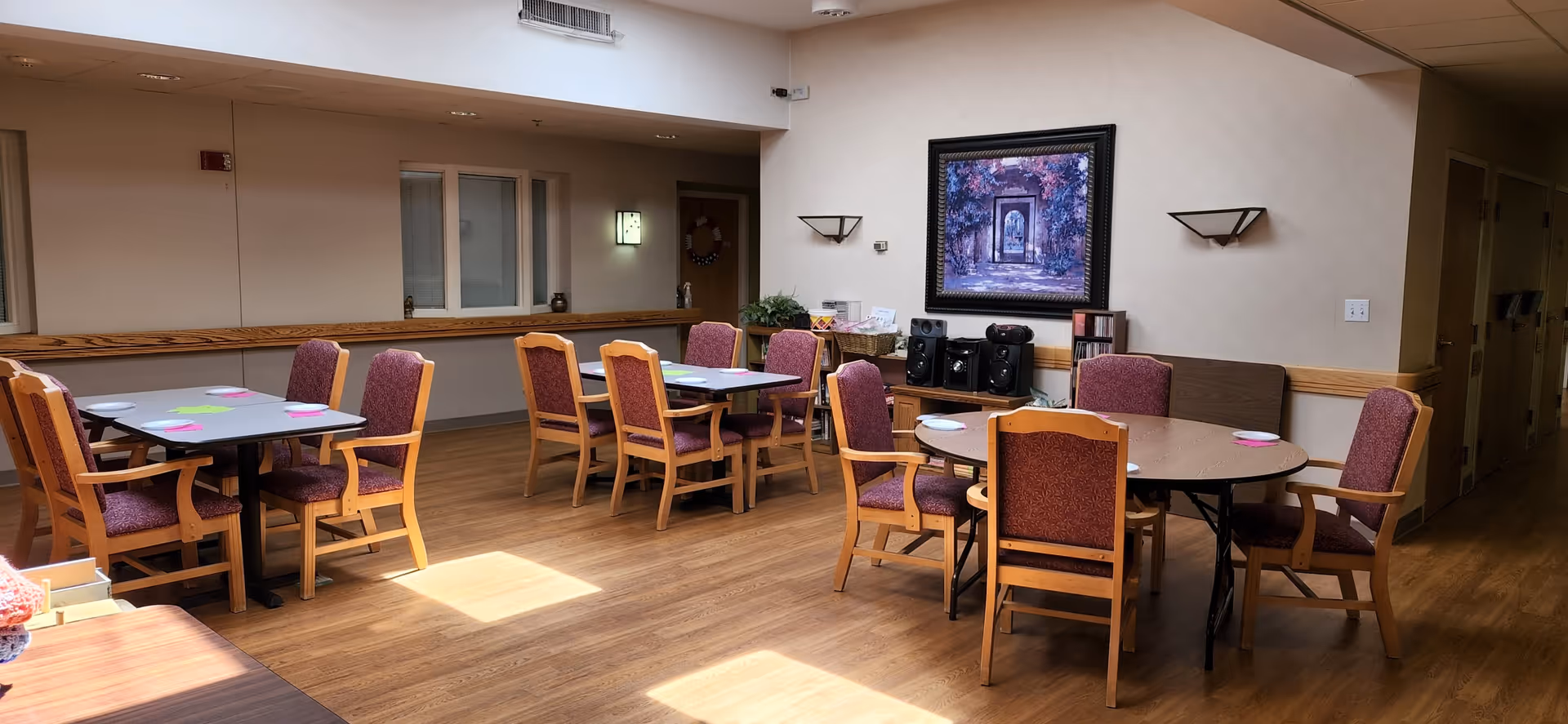 Sunlit common room with round and rectangular tables surrounded by upholstered wooden chairs and a framed painting on the wall.