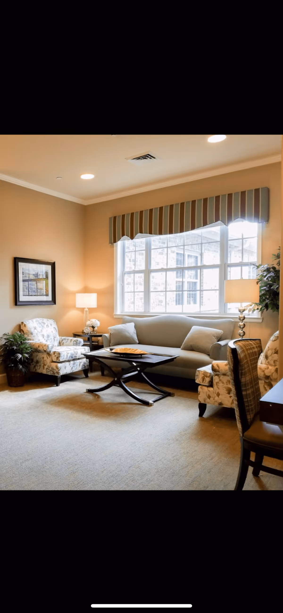 Cozy sitting room with a sofa, two patterned armchairs, a central coffee table, table lamps, and a large window with a striped valance.