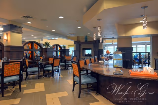 Interior view of a dining area with multiple tables and chairs featuring orange patterned upholstery. The space includes a large counter with bar stools, decorative lighting fixtures hanging from the ceiling, and a fireplace in the background. The room has large windows allowing natural light to enter.