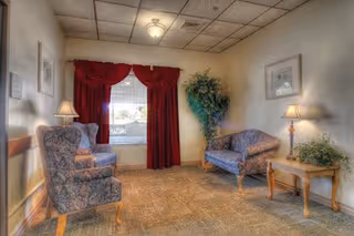 A cozy living room area with two patterned armchairs and a matching loveseat. There is a wooden side table with a lamp and a potted plant on it. A large window with red curtains and white blinds lets in natural light. The walls have framed artwork and the ceiling has a grid pattern with a central light fixture.