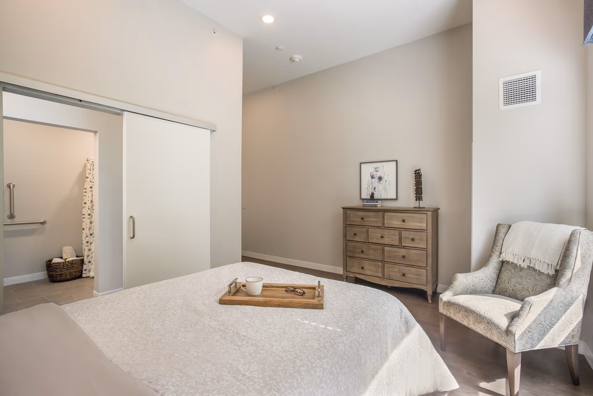 Bright furnished bedroom with a bed, wooden dresser, upholstered chair, and a sliding door opening to a bathroom.