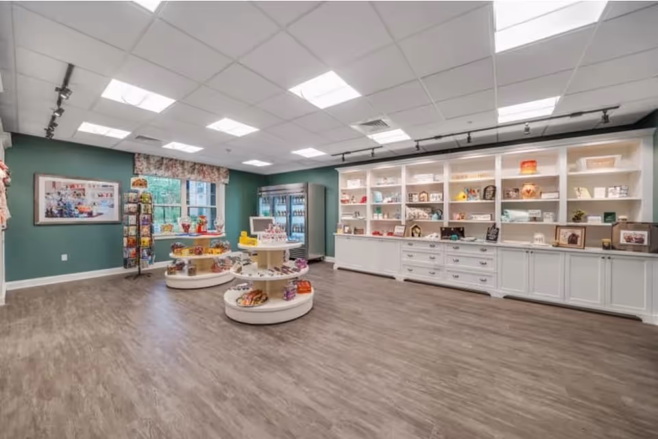 Interior view of a retail or gift shop area within a senior living facility. The room features wood-look flooring, green walls, and a white ceiling with recessed lighting. There are two round display tables in the center stocked with various items, a tall rotating rack with greeting cards near a window, and a large white shelving unit along one wall filled with decorative items and small merchandise. A refrigerated display case is visible in the back corner.