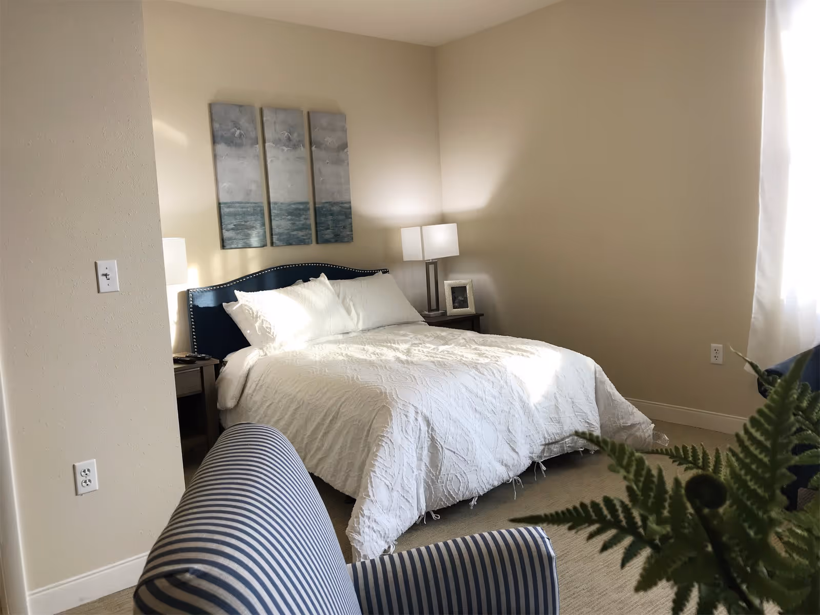 A bedroom with a neatly made bed featuring a white textured comforter and pillows. The bed has a dark blue upholstered headboard with nailhead trim. On either side of the bed are nightstands with lamps, one also holding a framed photo. Above the bed are three vertical panels of artwork depicting a cloudy sky over a body of water. A striped blue and white armchair is partially visible in the foreground, along with a green fern plant. Light streams in from a window with white curtains on the right side of the room.