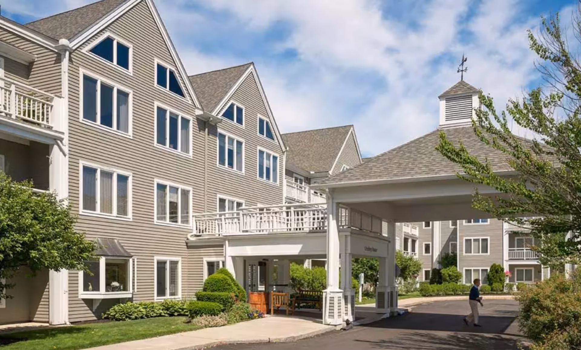Exterior view of a multi-story senior living facility with beige siding, multiple windows, and a covered entrance. There is a person walking on the driveway near the entrance, surrounded by greenery and trees under a partly cloudy sky.