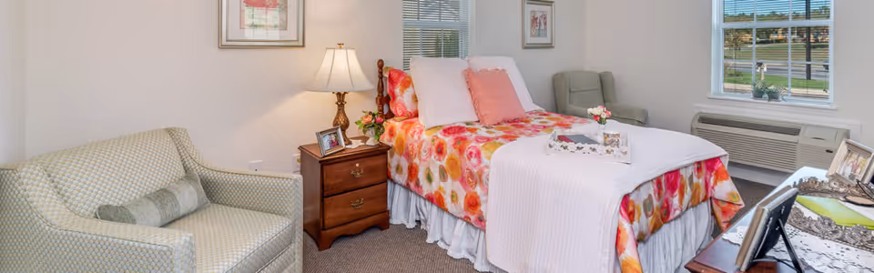 A bright and cozy bedroom featuring a single bed with floral bedding and pink pillows. Next to the bed is a wooden nightstand with a lamp, framed photos, and a small vase with flowers. There is a patterned armchair with a cylindrical pillow on the left side and another cushioned chair near the window. The room has two windows letting in natural light and an air conditioning unit below one window. A tray with a book and flowers is placed on the bed.