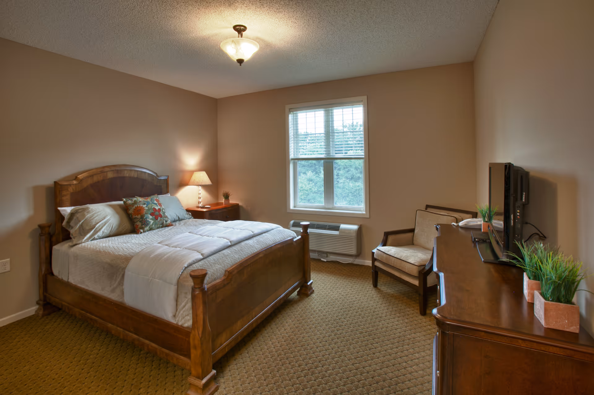 A cozy bedroom with a wooden bed frame, white bedding, and decorative pillows. There is a wooden nightstand with a lamp and a small plant next to the bed. A window with blinds lets in natural light, and below it is a wall-mounted air conditioning unit. Across from the bed is a wooden dresser with a flat-screen TV and two small potted plants. A cushioned armchair is placed beside the dresser. The room has beige walls and carpeted flooring.
