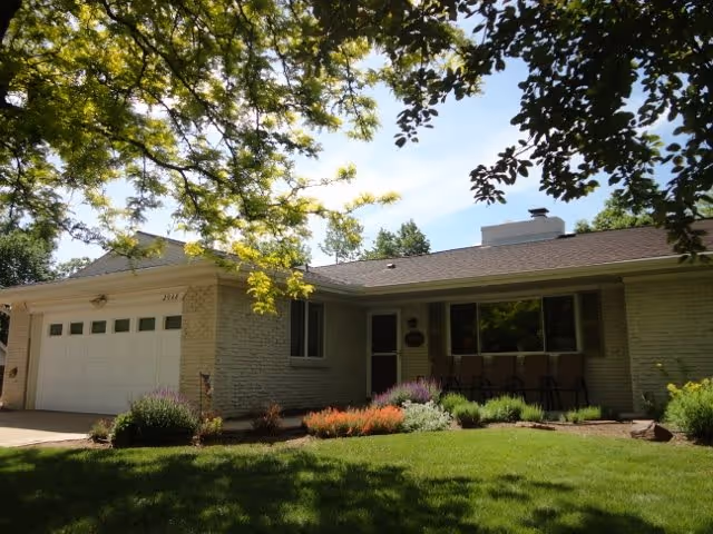 Exterior view of a single-story brick house with a garage on the left side, a front door, and a row of chairs on the porch. The house is surrounded by a well-maintained lawn and garden with various plants and flowers, and there are tree branches with green leaves partially framing the top of the image.