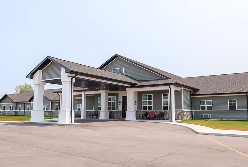 Exterior view of The Heartlands facility showing a single-story building with gray siding, white pillars, and a covered entrance. The building is surrounded by a paved driveway and some green lawn areas.