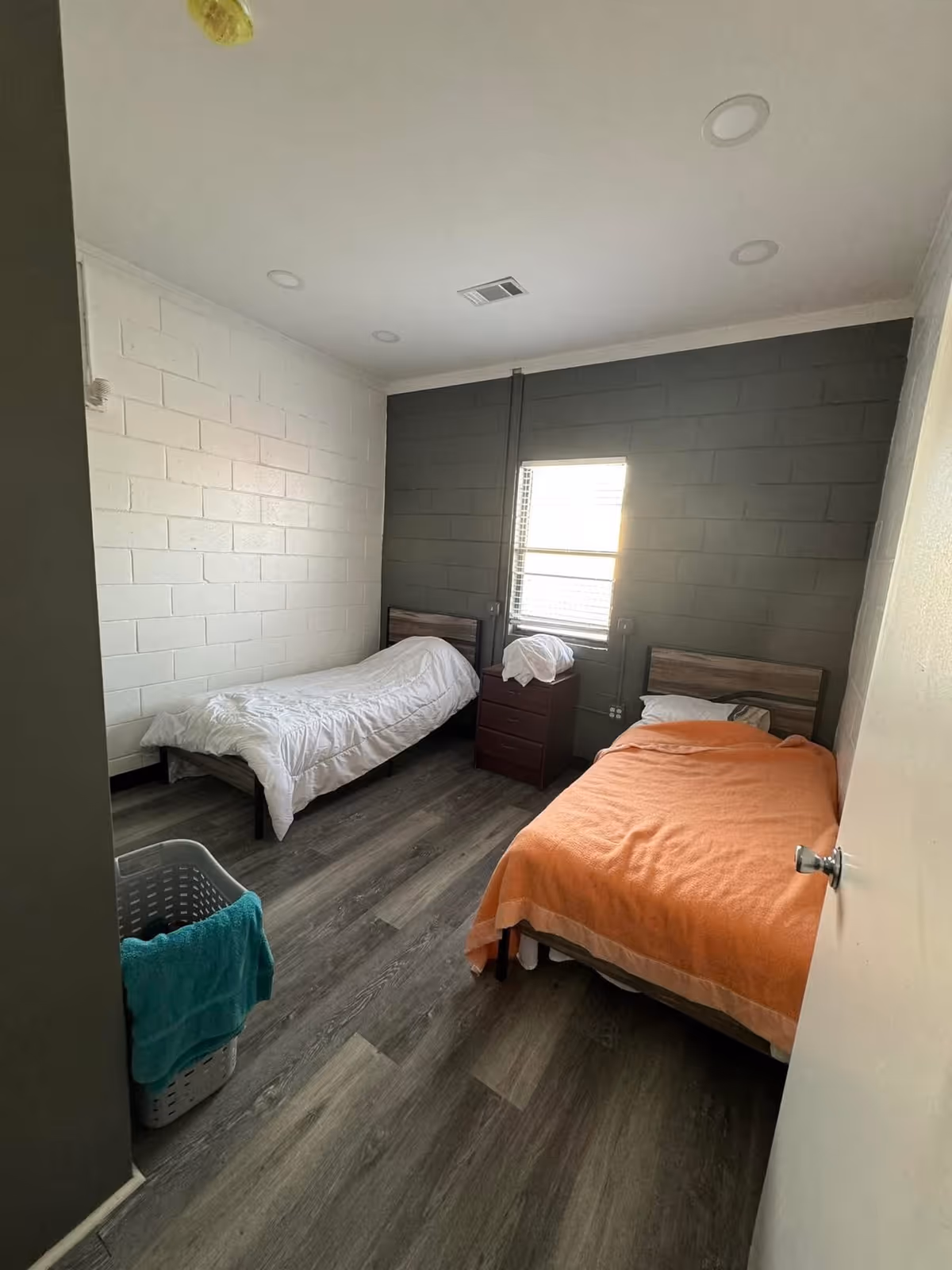 A small bedroom with two single beds, one with a white comforter and the other with an orange blanket. There is a window between the beds with blinds partially open, a small wooden nightstand with folded linens on top, and a laundry basket with a teal towel hanging on it. The walls are painted white and dark gray, and the floor has wood-like vinyl planks.