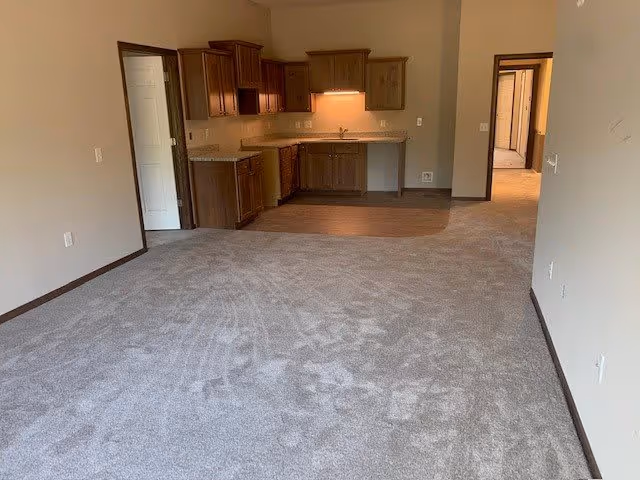 Spacious interior of an apartment featuring a carpeted living area and a kitchen with wooden cabinets and granite countertops.
