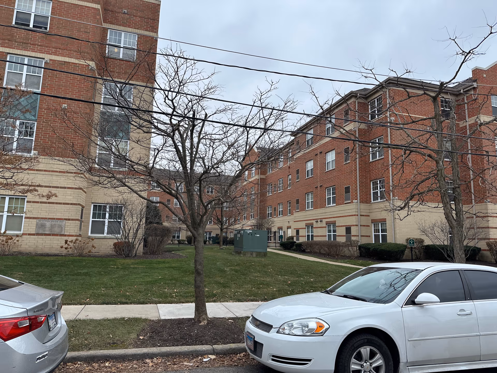 Exterior view of a brick multi-story residential building with leafless trees and parked cars under an overcast sky.