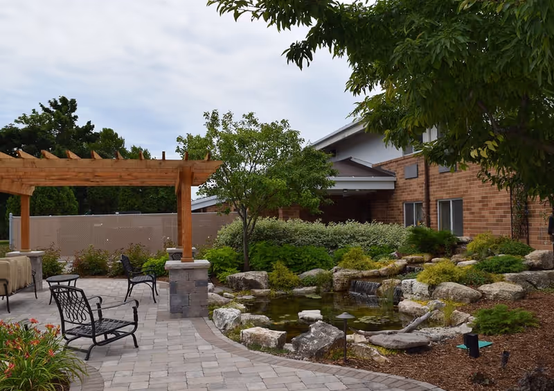 Outdoor patio area at Congregational Home featuring a paved walkway, black metal chairs, a wooden pergola, landscaped garden with rocks, a small pond with a waterfall, and trees and shrubs surrounding the space.