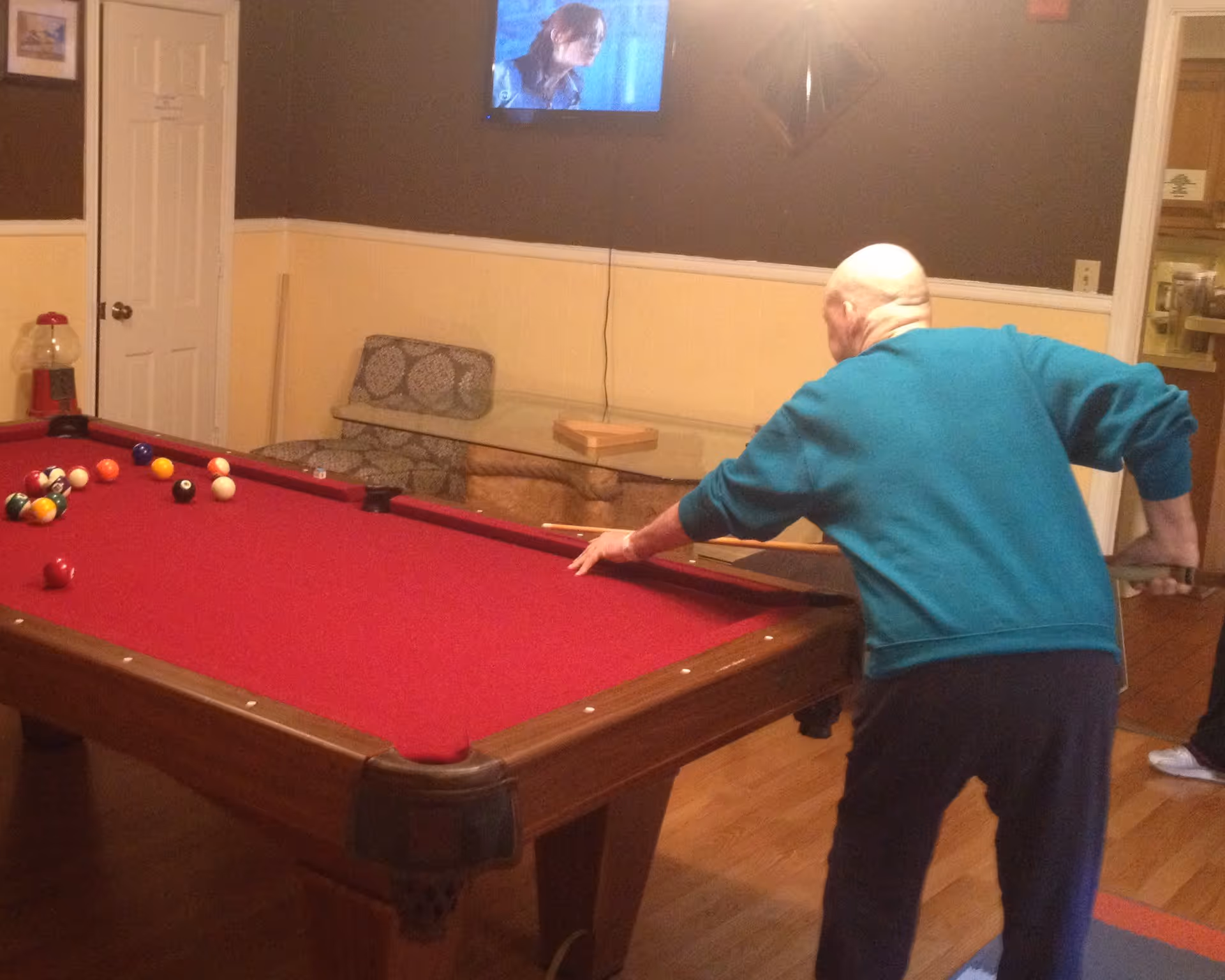 An older man leans over a red-felt pool table playing billiards in a cozy recreation room.