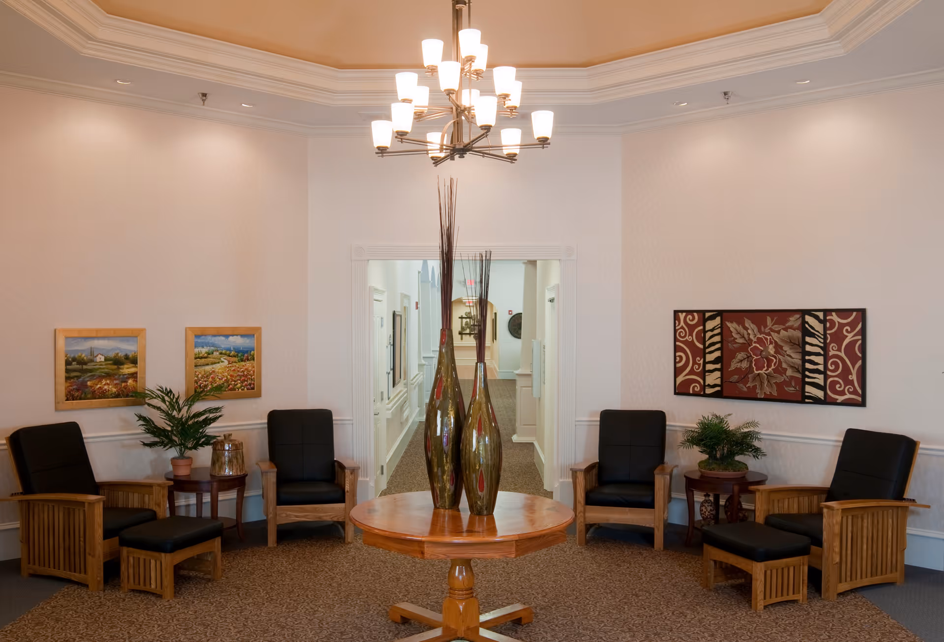 A cozy seating area in a senior living facility with four wooden armchairs featuring black cushions arranged around a round wooden table with two tall decorative vases. The walls are adorned with framed artwork and there are small side tables with potted plants. A chandelier hangs from the ceiling, and a hallway is visible in the background.