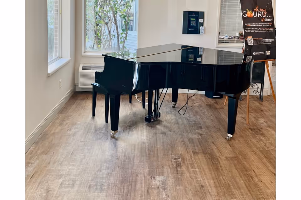 A black grand piano with a matching bench is placed on a wooden floor in a bright room with large windows. Outside greenery is visible through the windows. There is a white air conditioning unit beneath one window and a black signboard with text and a pumpkin graphic on the right side of the piano.