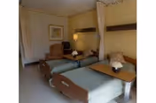 A room with two hospital-style beds each equipped with a wooden overbed table and a small flower arrangement. There is a comfortable armchair and a framed picture on the wall. The room has privacy curtains between the beds and soft yellow walls.