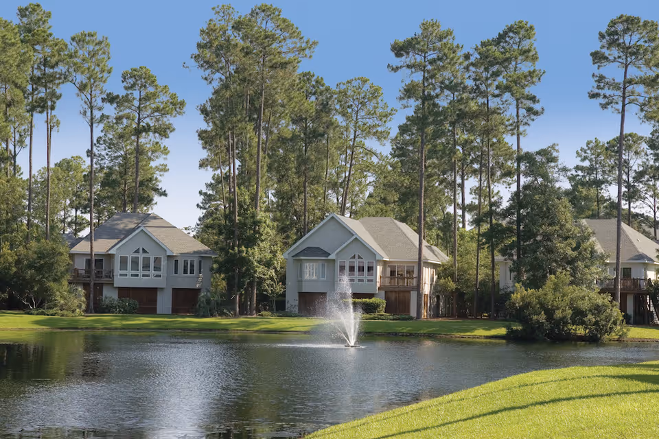 A serene outdoor scene at TidePointe, a Vi Community, featuring a small lake with a water fountain in the center, surrounded by green grass and tall pine trees. In the background, there are several two-story residential buildings with large windows and balconies.