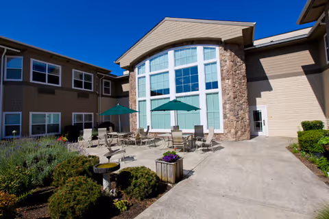 Outdoor patio area of a senior living facility with several tables and chairs under green umbrellas, surrounded by landscaped bushes and plants, adjacent to a building with large windows and stone accents.