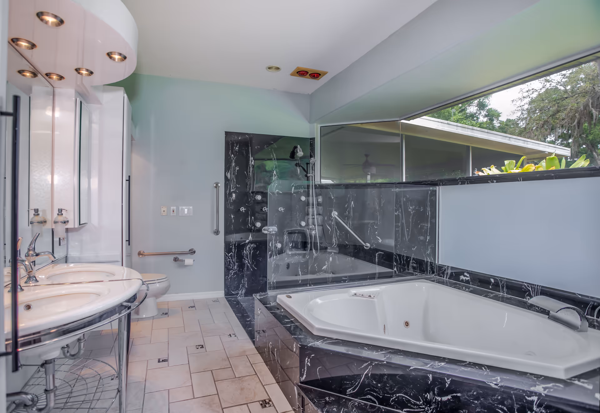 A modern bathroom featuring a large white bathtub with black marble surround, a glass shower enclosure with black marble walls, a double sink vanity with mirrors and overhead lights, a toilet with grab bars, and a large window providing natural light and a view of greenery outside.