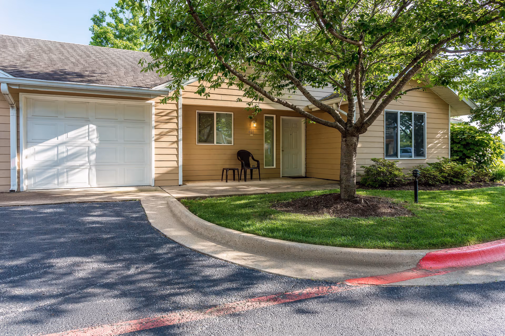 Exterior view of a single-story residential building with beige siding, a white garage door, a front porch with a black chair and small table, and a tree with green leaves in the front yard. The driveway and a red-painted curb are visible in the foreground.