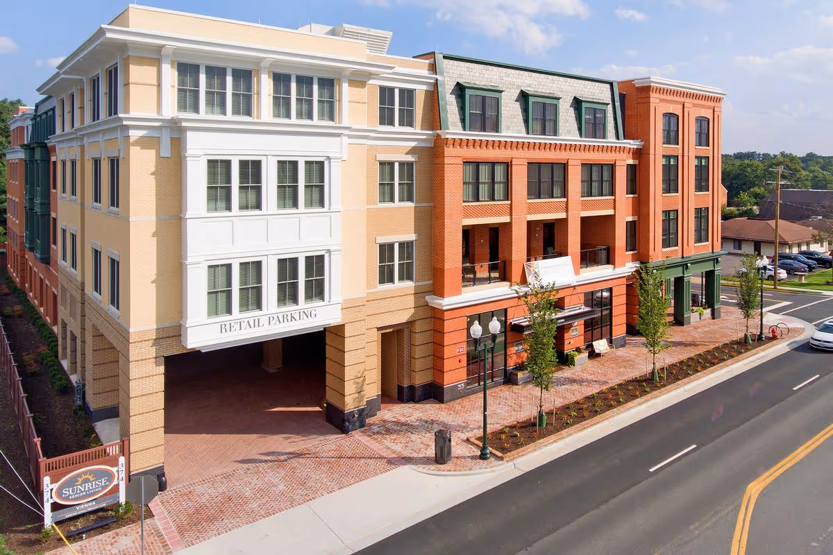 Exterior view of a multi-story building with retail parking underneath, a brick sidewalk with small trees, and a street with cars. A sign in front reads Sunrise of Vienna Senior Living.