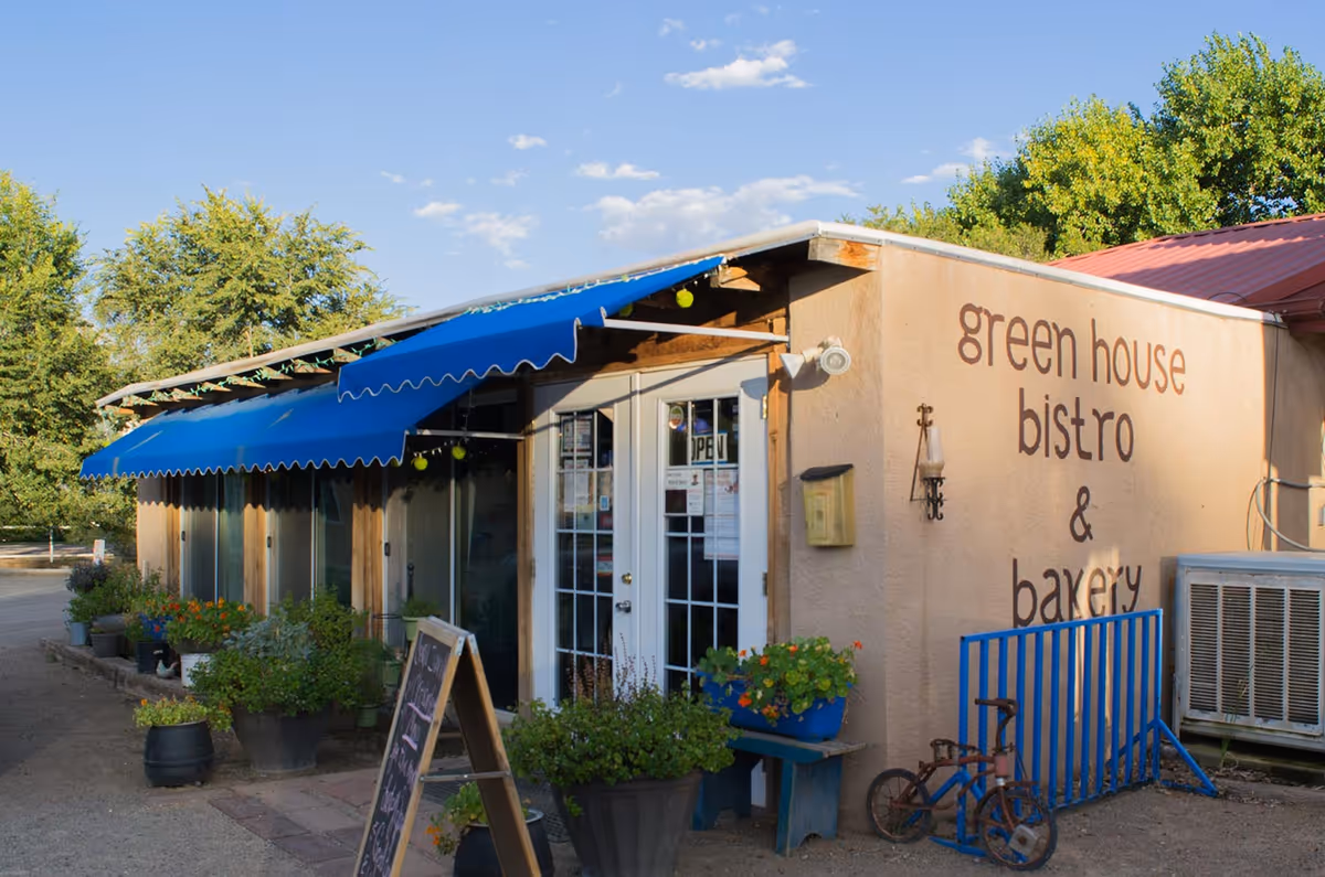 Exterior view of a small building with a blue awning and glass double doors. The building has a sign that reads 'green house bistro & bakery'. There are several potted plants and flowers outside, a chalkboard sign, and a small vintage tricycle near a blue metal railing. Trees and a clear sky are visible in the background.