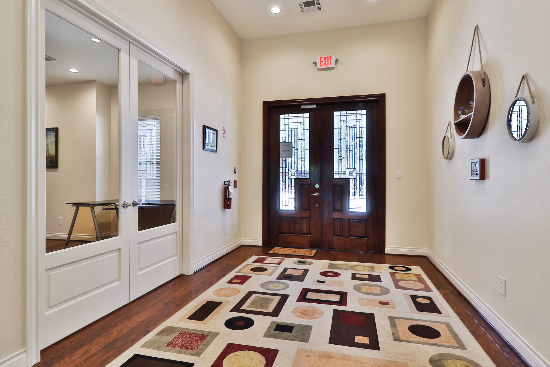 Entrance area of a facility with double wooden doors featuring decorative glass panels, a colorful geometric patterned rug on the wooden floor, white walls with round wall shelves and mirrors, and double glass doors on the left side leading to another room.