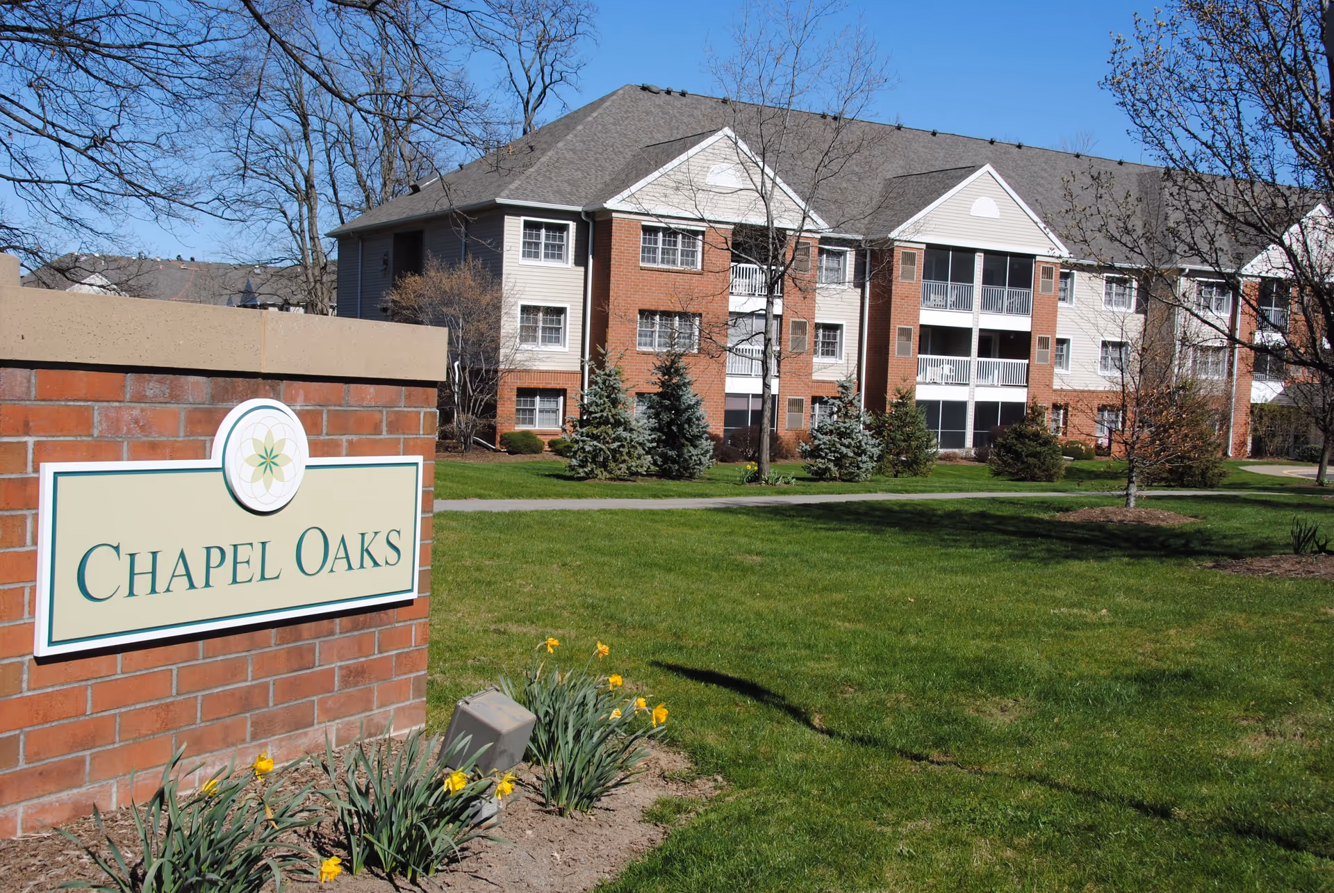 Exterior view of a senior living facility named Chapel Oaks with a brick sign in the foreground and a multi-story residential building with balconies in the background, surrounded by green grass, trees, and some yellow flowers under a clear blue sky.