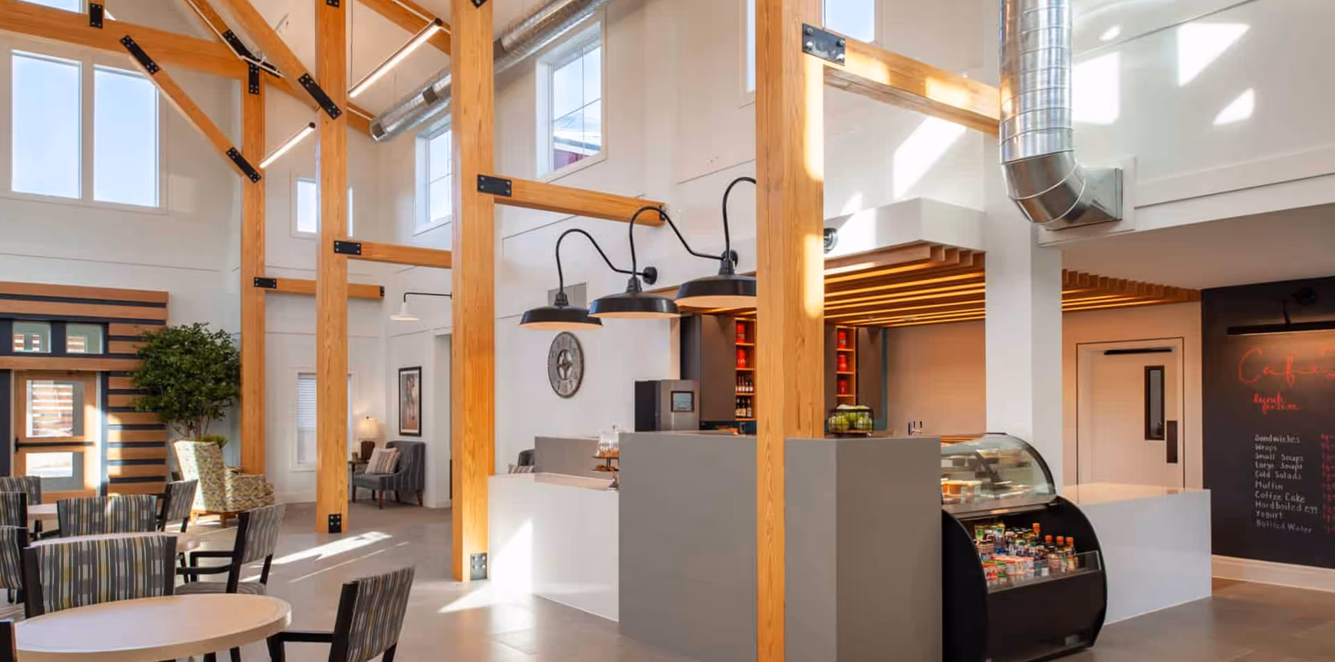 Bright open communal dining and café area with tall wooden beams, tables and chairs, and a service counter with a refrigerated display case.