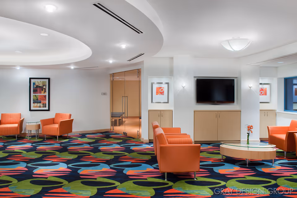 A modern lounge area with bright orange armchairs arranged around a round glass coffee table with a small flower vase. The room features a colorful carpet with abstract patterns, white walls, a flat-screen TV mounted on the wall, and framed artwork. There is a glass door leading to another room in the background.