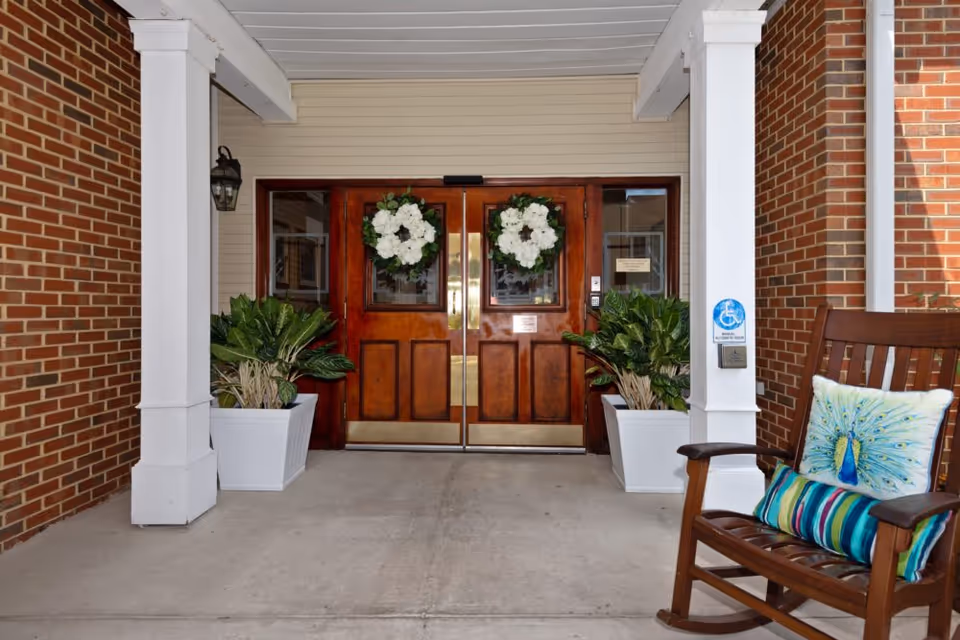 Entrance to a building with double wooden doors decorated with white floral wreaths. The entrance is framed by brick walls and white columns, with two large potted plants on either side of the doors. A wooden rocking chair with colorful cushions, including one with a peacock design, is placed to the right. A handicap accessibility sign and button are mounted on the right column.