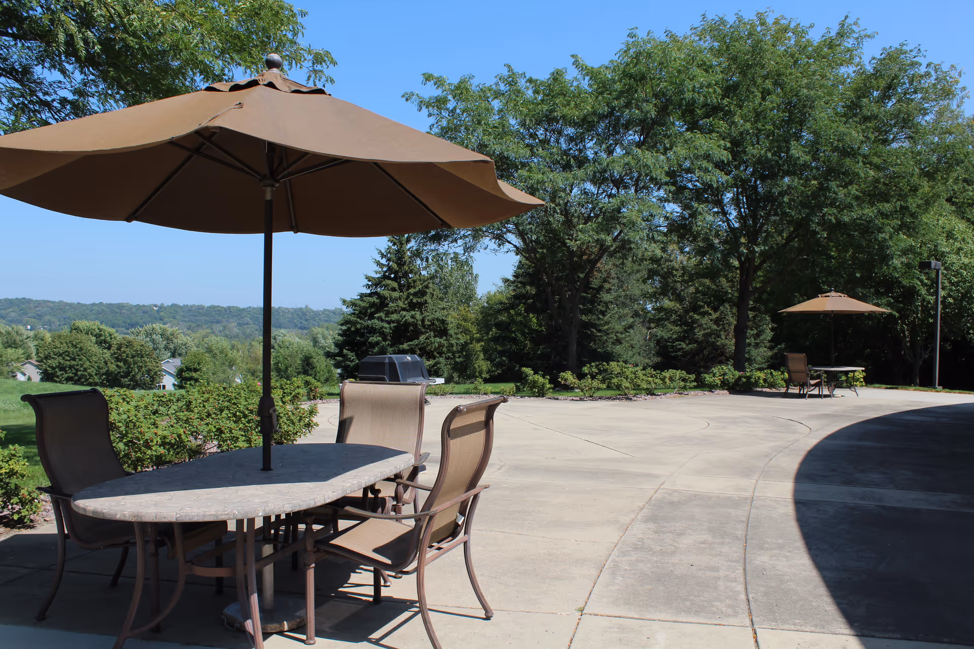 Outdoor patio area with two sets of tables and chairs, each shaded by large brown umbrellas. The patio is surrounded by greenery including trees and bushes, with a clear blue sky in the background.